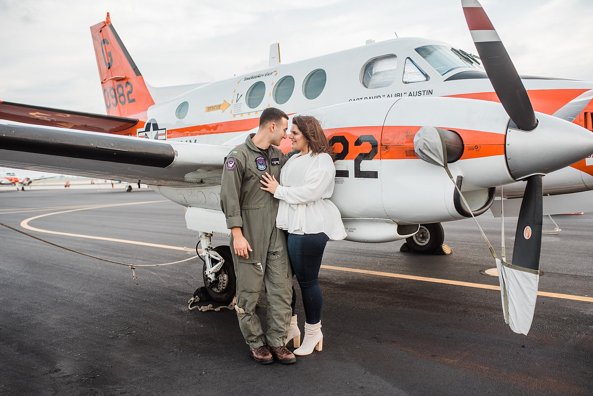 Navy Pilot in Green flight suit on flightline next to white plane with orange stripe with fiance in white top and blue jeans