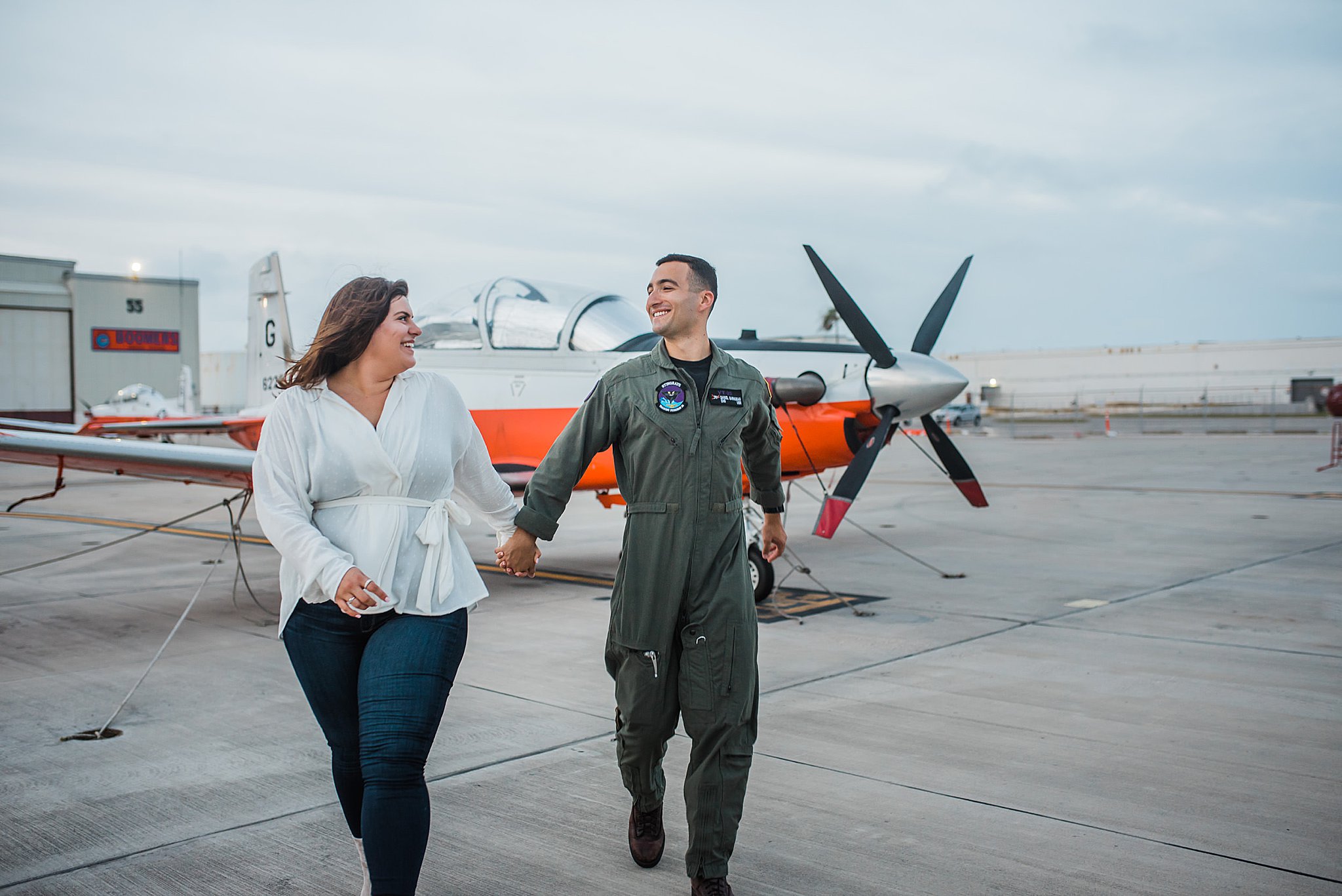 Navy Pilot in Green flight suit on flightline running in front of white plane with orange stripe with fiance in white top and blue jeans