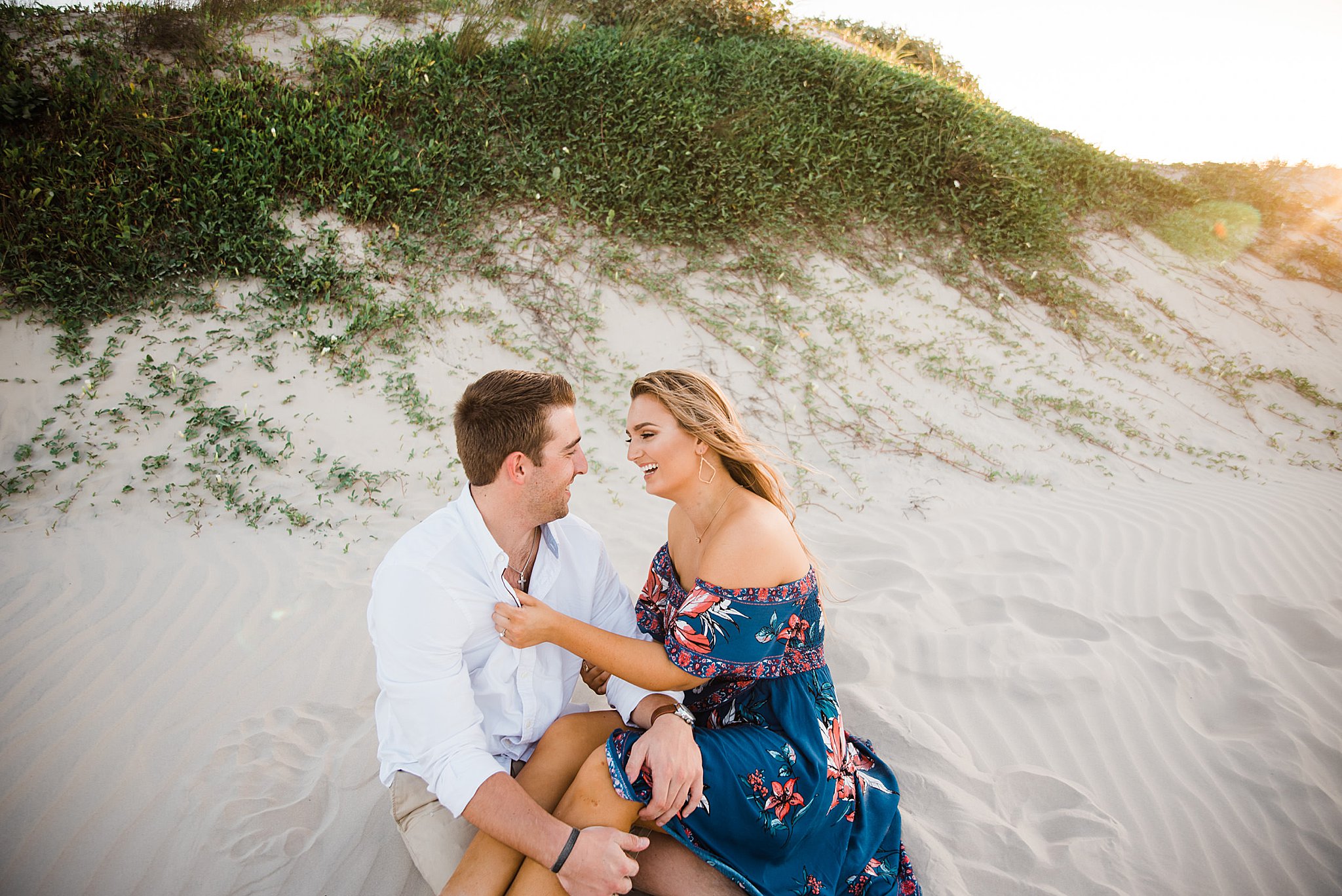 Man in white shirt and khaki short with woman in blue floral dress seated on the beach with greenery in background during north padre island engagement