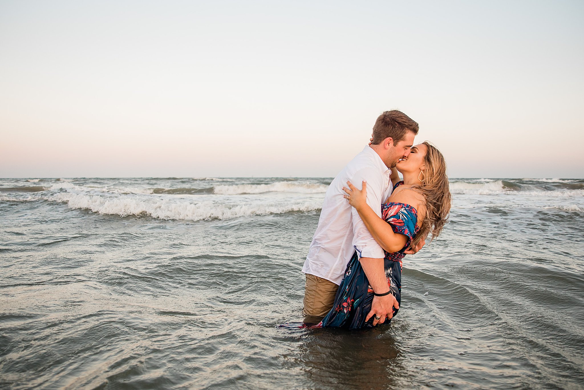 Man in white shirt kissing and embracing woman in blue floral dress waist deep in the ocean during north padre island engagement.