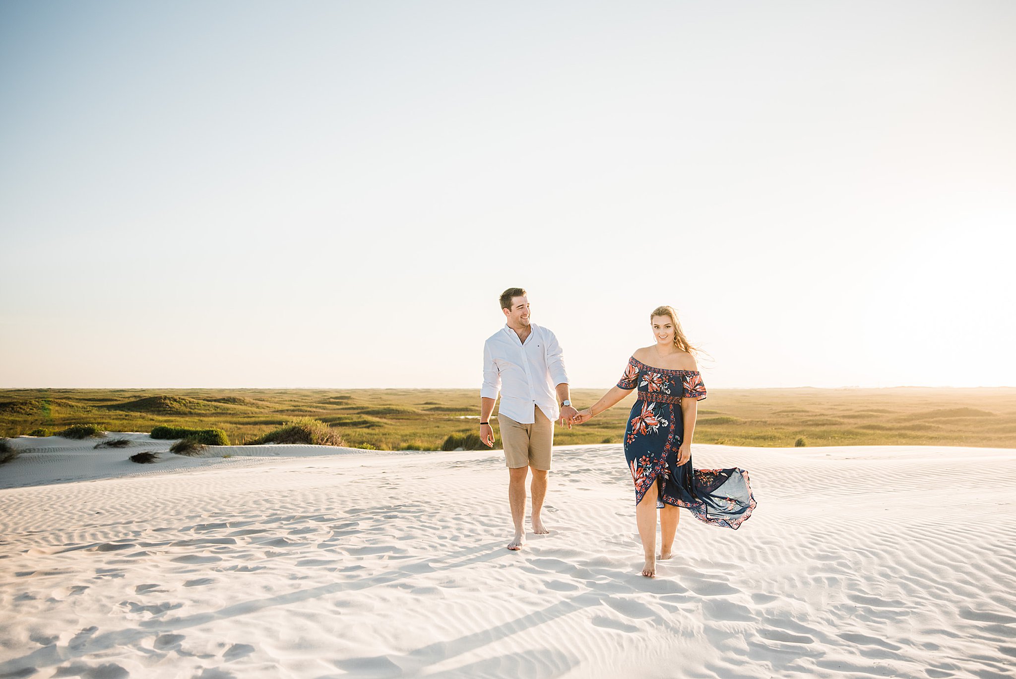 Man in white button up shirt holding hands and walking with woman in blue floral dress on beach during north padre island engagement